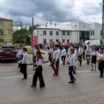 Orkiestra dęta maszeruje ulicą w czapkach i białych koszulach. W tle budynki i chmury. Flagi dodają kolorów.