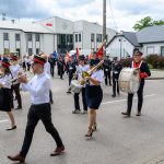 Orkiestra dęta w kolorowych mundurach maszeruje ulicą. W tle biały budynek, flaga Polski i chmury na niebie. Grają na trąbkach i bębnach.