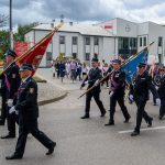 Strażacy w paradnych mundurach niosą flagi, idąc ulicą przed budynkiem z białą fasadą. W tle zauważyć można grupę ludzi. Niebo jest pochmurne.
