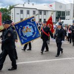 Grupa strażaków maszeruje ulicą, niosąc niebieskie i czerwone sztandary. Tło to białe budynki z flagą. Uczestnicy w ciemnych mundurach.