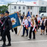 Parada na ulicy; ludzie w mundurach i młodzież z flagami. Tło z budynkami i drzewami, niebo pochmurne. Kolory jasne i kontrastowe.