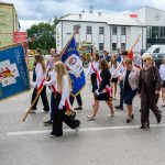 Grupa młodych ludzi maszeruje z niebieskimi i białymi chorągwiami na ulicy. Ubrani są w białe koszule z czerwonymi szarfami. W tle znajduje się budynek i zielone drzewa.
