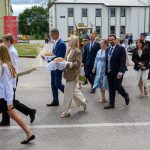 Grupa ludzi w eleganckich ubraniach idzie ulicą przy budynkach, trzymając chleb na białych serwetkach. Flaga z orłem i czerwonym tłem. Otoczenie jest zielone z chmurami na niebie.