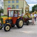 Na zdjęciu widoczny jest kolorowy traktor z przyczepą ozdobioną plonami i słomianymi wieńcami. W tle stoi żółto-zielony budynek, a na ulicy widoczna jest grupa osób. Sceneria przypomina tradycyjny festiwal dożynkowy.