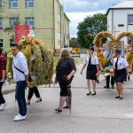 Grupa ludzi niesie ozdobne wieńce z zboża, na jednym obraz Matki Boskiej. Tło to budynki i drzewa. Atmosfera jest uroczysta.