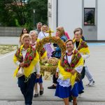Grupa kobiet w tradycyjnych, kolorowych strojach niesie dekoracyjny wieniec. Tło to budynek i drzewa. Atmosfera świąteczna.
