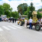 Parada na ulicy: traktor ciągnący kolorowo udekorowaną przyczepę. W tle uczestnicy niosą flagi. Zielona sceneria dookoła.