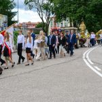Uroczysta procesja idzie ulicą, ludzie ubrani formalnie, wśród zieleni i miejskich budynków. Tło stanowią drzewa i budynki. Kolory dominujące to zieleń, biel oraz czerwony akcent flag.