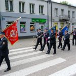 Grupa strażaków w mundurach maszeruje z kolorowymi sztandarami na przejściu dla pieszych. Budynki w tle są szare, a pogoda pochmurna. Na banerach dominuje kolor czerwony i niebieski.