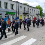 Parada strażaków w mundurach maszerujących ulicą z flagami. Tło stanowią budynki i drzewa. Kolory to głównie niebieski i czerwony.