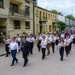 Orkiestra maszeruje ulicą w kolorowych mundurach, grając instrumenty. Tło stanowią budynki i drzewka. Po prawej stronie jest chodnik z kostki.