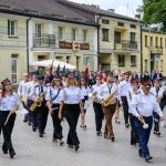 Orkiestra dęta maszeruje ulicą, muzycy w białych koszulach i czapkach. W tle są kremowe budynki. Zieleń po prawej stronie.