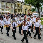 Parada orkiestry dętej w białych koszulach i czapkach na ulicy. Tło to budynki z szyldami i zielone drzewa. Zanurzeni w muzyce, maszerują.