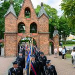 Parada strażaków w czarnych mundurach przed ceglaną dzwonnicą. Kolory to czerwień cegieł i zieleń drzew. Ludzie oglądają z boku.