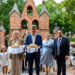 Cztery osoby w eleganckich strojach trzymają chleby na białych serwetkach przed ceglanym budynkiem z dzwonami. W tle zielone drzewa i krzyż na szczycie. Obecni są również ludzie w tle, co wskazuje na uroczystość.