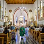 Wnętrze kościoła z drewnianym sufitem i żyrandolami. Ksiądz w zielonej szacie przechodzi przez nawy pełną ludzi. Tło zdobią witraże i obrazy religijne.