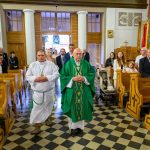 Procesja w kościele; ksiądz w zielonej szacie i ministrant w białej idą środkiem między ławkami. Drewniane ławki, kolorowa posadzka. Uczestnicy stoją po bokach, w tle witraże.