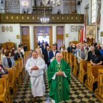 W jasnym kościele ksiądz w zielonej szacie prowadzi procesję. Na ławkach siedzą wierni w strojach ludowych. Ściany zdobią dekoracje i teksty biblijne.