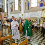 W kościele, procesja z duchownym w zielonym ornacie. W tle osoby siedzące, orkiestra, freski i żyrandol. Pastelowe ściany.