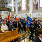 Procesja strażaków w mundurach z flagami w kościele pełnym ludzi. Ściany zdobią freski i napisy, a wnętrze jest jasne.