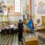 Kościół z wiernymi podczas ceremonii, orkiestra dęta na tle witraży. Dominuje kolor biały i złoty we wnętrzu. Procesja z flagą strażacką.