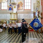 Uroczystość w jasnym kościele z orkiestrą i sztandarem straży pożarnej. Na przodzie mężczyzna trzyma chleb w białych serwetkach. Ściany zdobią religijne malowidła i złote detale.