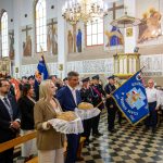 Na obrazie widoczna jest uroczystość w kościele z ludźmi niosącymi chleby. Tło zdobią kolorowe freski i flagi strażackie. Dominują kolory biały, niebieski i złoty.