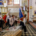Grupa uczniów z flagami wchodzi do kościoła. Tło ozdobione malowidłami i złotymi elementami. Podłoga w czarno-białą szachownicę.