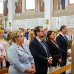 Zdjęcie przedstawia grupę ludzi w kościele podczas nabożeństwa. Widać kolorowe witraże i malowidła ścienne w tle. Osoby są elegancko ubrane i stoją w rzędzie.