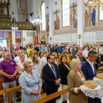 Wnętrze kościoła pełne ludzi uczestniczących w uroczystości. Dominują kolory białe i brązowe, mury zdobią barwne freski. Na pierwszym planie para trzyma ozdobne chleby.