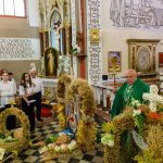 Ceremonia kościelna z kapłanem w zielonym ornacie. Otoczenie ozdobione wieńcami z kłosków i kwiatów. W tle biało-złote elementy ołtarza.
