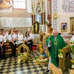 Kapłan w zielonym ornacie błogosławi wieńce dożynkowe w kościele. Obok grupa osób w białych koszulach. Tło ozdobione jest religijnymi obrazami i rzeźbami.
