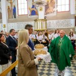 Kapłan w zielonych szatach błogosławi chleb w kościele, otoczony wiernymi i chórem. Wnętrze zdobią freski i witraże.