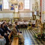 Ludzie w kościele na uroczystości, siedzą na ławkach. Dekoracje z kwiatów i zboża. Tło z freskami i witrażami.