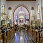 Wnętrze kościoła z tłumem wiernych. Kolorowe witraże i freski zdobią ściany. W centrum widoczny jest bogato zdobiony ołtarz.