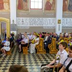 Obraz przedstawia ludzi siedzących w kościele na drewnianych ławkach. W tle są malowidła ścienne i cytaty. Kolorystyka jest jasna, dominują beże i brązy.