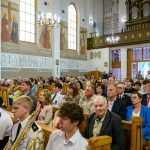 Ludzie siedzą w kościele ozdobionym freskami i witrażami. Jasne wnętrze z drewnianymi ławkami i złotymi elementami. Na ścianach wiszą obrazy religijne.