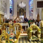 Kościół wewnątrz z ozdobnymi wieńcami zbożowymi i obrazem Maryi. W tle ołtarz, witraże, ksiądz przy ambonie. Kolory jasne, złote i zielone.