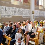 Wnętrze kościoła wypełnione ludźmi w eleganckich ubraniach. Na ścianach są malowidła i napisy religijne. Kilka osób ma wianki z kwiatów.