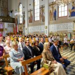 Wnętrze kościoła z wiernymi na drewnianych ławkach, jasne ściany z malowidłami religijnymi i witraże. Dekoracje w złotej kolorystyce. Na suficie wiszą żyrandole.