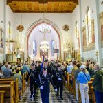 Grupa strażaków w mundurach maszeruje przez kościół. Wnętrze jest jasne z drewnianym stropem. W tle widać ołtarz i witraże.