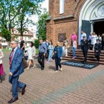 Grupa ludzi wychodzi z kościoła z ceglaną fasadą. Tło to drzewa i budynki. Kolory są jasne, z granatowymi i różowymi akcentami.