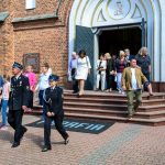 Grupa ludzi wychodzi z ceglanego kościoła z białym wykończeniem, na schodach widoczni strażacy. Słoneczny dzień z zielenią w tle.