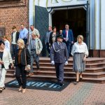 Grupa ludzi opuszcza ceglaną budowlę z dużymi drzwiami. Ubrani są elegancko, widać mundur. Tło to czerwona cegła i schody.