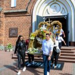 Ludzie wychodzą z kościoła z ozdobnym wieńcem w kształcie figury zboża. Ceglana fasada i otwarte drzwi w tle. Kolory bieli i zieleni.