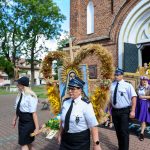 Ludzie w mundurach niosą wieniec z wizerunkiem Matki Boskiej przed ceglaną świątynią. Zieleń drzew i niebieskie niebo wokół. Dominują kolory bieli, złota i brązu.