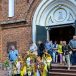 Grupa ludzi opuszcza ceglaną świątynię z ozdobną bramą. Kobiety w kolorowych strojach niosą żniwne wianki. Tło z cegły i białymi detalami.