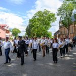 Orkiestra strażacka maszeruje ulicą w piękny, słoneczny dzień. Czerwono-czarne mundury kontrastują z zielenią drzew. W tle widoczny jest kościół.