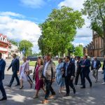 Grupa ludzi w eleganckich strojach idzie ulicą. Tło to zielone drzewa i ceglana zabudowa. Niebo jest błękitne z białymi chmurami.