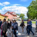 Grupa osób w formalnym ubraniu spaceruje po mieście. Tło to jasne budynki i zielone drzewa pod niebieskim niebem. Atmosfera spotkania w pogodny dzień.